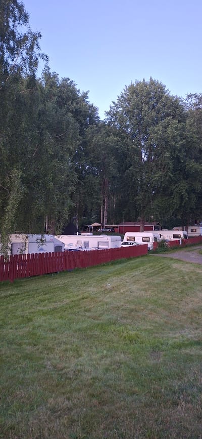 The image depicts a scene in Salpakangas, Finland, featuring a row of parked caravans behind a red wooden fence. Lush green grass covers the foreground, with a slight incline. Tall, mature trees with dense foliage dominate the background, casting shadows and creating a natural backdrop. The sky is clear and a light blue, suggesting daytime. Several white caravans are visible, lined up parallel to the fence. Some of these caravans have windows and recreational vehicle features. Beyond the caravans, glimpses of buildings with red roofs and covered structures can be seen, possibly part of a campground or residential area. The overall impression is a tranquil outdoor setting, likely a caravan park or a residential area with ample green space and trees.