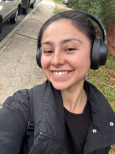 A person is taking a selfie outdoors. The person is wearing black headphones and a black jacket. A row of cars are parked along the road, and the backdrop consists of greenery. The location is in Bassendean, Australia, judging from metadata not shown.
