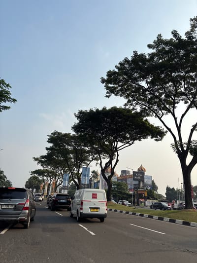 The image shows a street scene in Pakulonan Barat, Indonesia, likely during the daytime with a clear sky. A line of vehicles is on the road, with a silver SUV in the foreground on the left, followed by a white van, and other cars further down the road. Trees line the side of the road, casting shadows. In the background, there are several buildings with signs, including "HANDAL FURNIT", "KING KOIL", and "THERAPEDIC". Banners with "ONE DISTRICT TWO ICONS" are also visible. The road has clear lane markings.