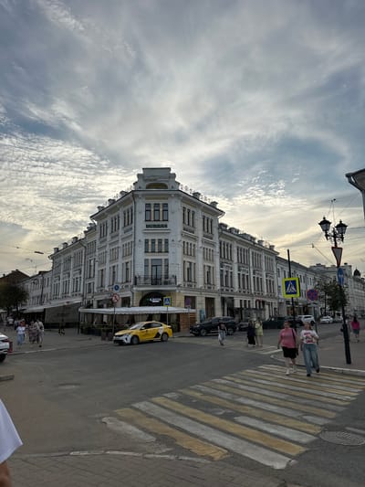 The image depicts a street scene in Putyatino (Semivragovskaya S/A), Russia, featuring a large, ornate, multi-story building on the right side of the frame. The building is white with detailed architectural elements, including decorative moldings, arched windows, and a prominent cornice. A yellow taxi is parked near the building, and several pedestrians are walking on the sidewalk and crossing a street at a marked crosswalk. The street is paved, and there are traffic lights and street lamps visible. The sky is partly cloudy, with a mix of white and gray clouds. The overall impression is that of a typical urban environment on a cloudy day.