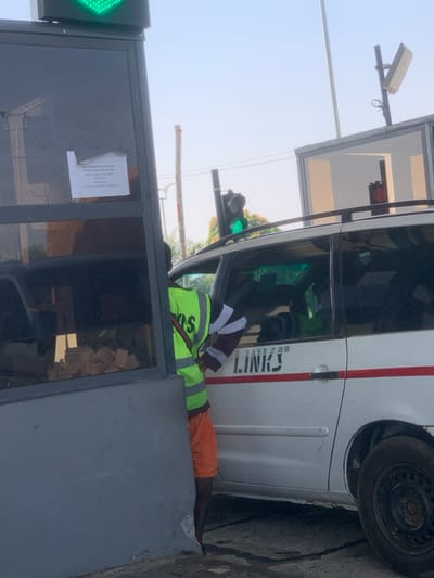 This image depicts a checkpoint or toll plaza in Kubare, Nigeria. A male attendant is positioned between a dark-paneled booth structure and the driver's side of a white multi-purpose vehicle. The attendant wears a neon green reflective vest with "POS" visible on the back, orange shorts, and a long-sleeved shirt with black and white striped sleeves; his right arm is bent, extended towards the vehicle's interior. The white vehicle, possibly a minivan, displays "LINKS" in black capital letters and a horizontal red stripe on its side. Its driver's side front window is partially open.
Above the scene, a large green traffic light indicates an open lane, while a smaller red traffic light is visible further down the lane, above the vehicle. A white rectangular sensor or antenna is mounted on a pole above the vehicle. Taped to the interior of the booth's dark glass window is a white sign with black text, partially visible, stating: "CASH PAYMENT NOT ALLOWED. PAYMENT METHOD INCLUDED. DON'T CREATE TRAFFIC. TRANSIT. THIS LANE IS FOR ELECTRONIC PAYMENT." The sky is clear, and a utility pole stands in the background.