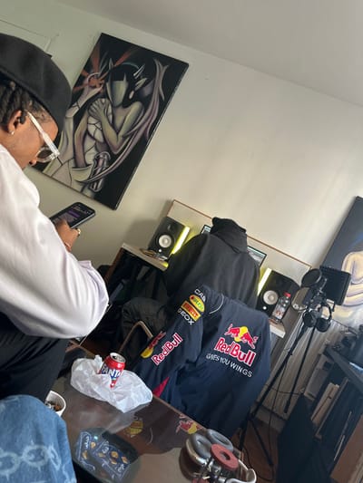 An indoor scene in Bondy, France, depicts two individuals in what appears to be a home studio or creative space. In the foreground to the left, a person with dark skin, short dreadlocks, and wearing a black cap and clear-framed glasses holds a black smartphone with both hands, looking down at its screen. They are dressed in a light-colored long-sleeved shirt.

Behind this individual, centered in the mid-ground, a second person is seated with their back to the viewer, wearing a dark hooded garment. This person is positioned at a desk, actively engaged with a computer screen, flanked by two studio monitor speakers with illuminated yellow cones. To the right of the desk, a microphone on a tripod stand, equipped with a pop filter and a pair of over-ear headphones, is present.

A dark blue racing jacket with prominent Red Bull branding and sponsor logos (Castrol, Pirelli, Geox, Rauch) is draped over a chair between the two individuals. On a glass-top coffee table in the immediate foreground, an orange Fanta can rests inside a clear plastic bag. Nearby, a small white bowl contains cigarette butts, and a dark-colored object with circular components, possibly a grinder, is also visible. Two small blue packages are on the table's surface.

The walls are painted white and display two distinct art pieces: on the left, a vertical painting depicts a stylized, dark-winged figure with horns in shades of purple, grey, and black; a second, partially visible painting with lighter tones hangs on the right wall. A radiator is visible along the lower right wall.