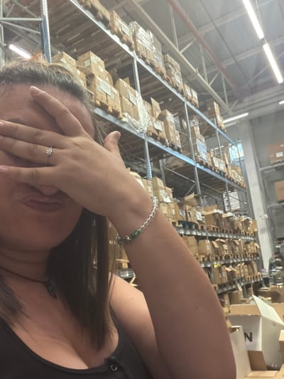 The image shows a woman covering her face with her hand in a warehouse. She is wearing a dark top and a bracelet on her wrist. A ring with a flower design is visible on her finger. The background features tall shelving units filled with cardboard boxes, indicative of a storage or distribution facility. The lighting suggests it is daytime, with fluorescent lights illuminating the space. The overall scene conveys a sense of being overwhelmed or tired, given the woman's gesture. The location is Gara Elin Pelin, Bulgaria. There is visible text on one of the boxes that reads "N".
