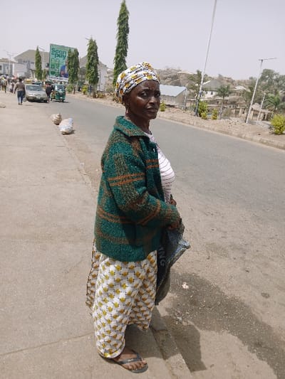 An adult female subject is depicted standing on a concrete sidewalk, partially facing the viewer while looking slightly to her right. She wears a yellow, white, and black patterned headwrap, a green and brown striped knitted jacket over a white and pink striped top, and white pants with a repetitive yellow pattern. Black flip-flops are on her feet. She holds a blue and white patterned woven bag, partially visible in her left hand. The sidewalk shows cracks and discolored patches.

Behind the subject, a paved road stretches into the distance, with a lighter-colored shoulder on the right. Several vehicles are visible on the road to the left, including a light-colored sedan and a green three-wheeled auto-rickshaw (tuk-tuk). Further down the road, two individuals walk on a sidewalk parallel to the subject. Two sacks are present on the ground near the roadside. Tall, slender green trees line the left side of the road. A large green billboard with the text "Scan to pay" and images of mobile phones is visible on the left. Utility poles are present on both sides of the road.

The background reveals scattered buildings and rocky, arid terrain, possibly a hillside, under a hazy, light-colored sky, indicating a bright, possibly dusty, outdoor environment. The scene is located in the city of Kadoma, Zimbabwe.