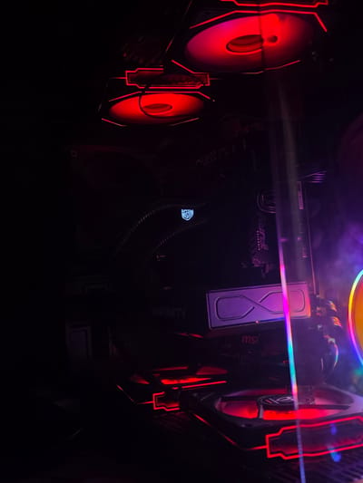 The image shows the interior of a computer case, illuminated by red and purple LED lights. Components like fans, a graphics card with "INFINITY" branding, and RAM sticks are visible. The scene is dark, with the lights creating a futuristic and vibrant atmosphere. There is no indication of location in Aleksandrovo, Bulgaria.