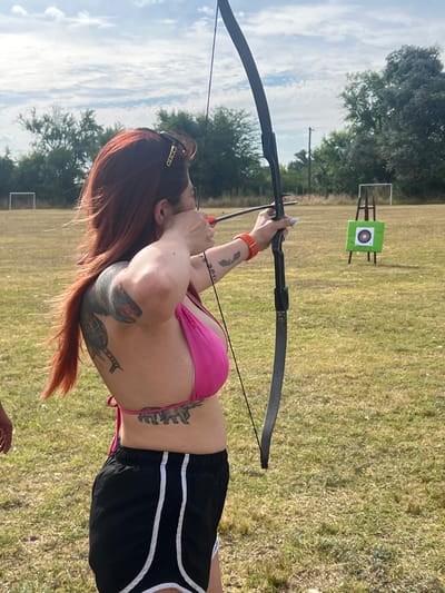 A person stands outdoors in a field, drawing back an arrow on a recurve bow. The individual is wearing a pink bikini top and black shorts with white stripes. Visible tattoos adorn their arm and torso. An archery target stands in the background, a green square with a bullseye. Trees and a cloudy sky form the backdrop to the scene.