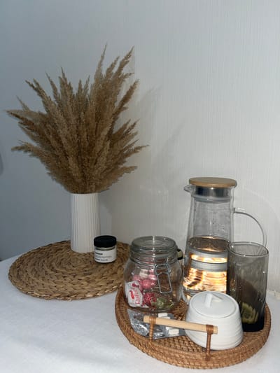 The image displays an indoor still life arrangement, likely on a table or counter, against a light-colored, subtly textured wall. The scene is presented with soft, diffused lighting, suggesting daytime.

On the left side, a large, fluffy bouquet of dried pampas grass or similar feathery reeds stands upright in a white, vertically ribbed ceramic vase. Beneath the vase is a round, woven placemat or coaster made of natural fibers.

To the right, a circular woven tray holds several items. These include a clear glass pitcher or carafe with a wooden lid and a metallic band, and next to it, another tall clear glass. A larger clear glass jar with a metal clip-top lid contains individually wrapped sweets, with the brand "Raffaello" clearly visible on one of the wrappers. Adjacent to this jar is a white ceramic jar with a lid, possibly for sugar or another condiment. A small wooden stick or scoop, secured by thin cord, is attached to the rim of the woven tray near the ceramic jar.

Between the vase and the tray, a small black-capped jar, likely a candle or diffuser, is visible. Visible text on its label reads "CHMB460H," "COLLECTION," and smaller text indicating "PARFUM / 200 g / 7.05 oz."

The overall aesthetic features natural textures and neutral colors, creating a serene and organized display. The items are arranged decoratively, typical of a home setting in Анкудиновка, Russia.