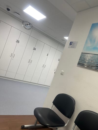 This image, captured from an elevated, angled perspective, depicts a clean, modern interior space, likely a waiting area or changing room in Herne, Germany. No people are visible.
The scene features a long wall of white, numbered lockers extending from the mid-ground into the background on the left side. Locker numbers "32," "33," "34," and "35" are clearly visible, each locker equipped with a silver handle and a keyhole. Above the lockers, a convex security mirror is mounted on the wall. Further left, near the ceiling, a small, black dome security camera is installed. The ceiling consists of white square panels with integrated rectangular fluorescent lights, one of which glows brightly.
On the right side of the image, three black, padded chairs with silver metal frames are arranged, suggesting a waiting area. The floor beneath these chairs is made of wood, transitioning to a light blue/grey speckled material covering the rest of the visible floor space.
The textured light-colored walls are adorned with a large rectangular picture depicting a serene landscape of a calm body of water under a light blue sky. Below this artwork, a white rectangular sign with a blue camera icon is affixed. Visible text on the sign includes the German word "Achtung" (Attention/Caution) and partially visible "Videoüberwachung" (video surveillance), indicating the presence of security cameras. A small, white, rectangular object, possibly a sensor or alarm, is also mounted on the wall.
The combination of lockers, waiting chairs, and surveillance equipment suggests a functional, semi-public environment such as a clinic, gym, or public facility.