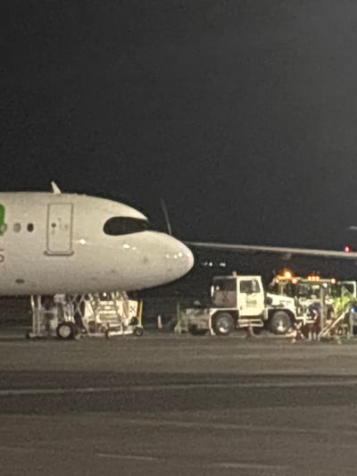 From an aerial perspective, the nose of a white aircraft is positioned adjacent to a dark tarmac. Next to the aircraft on the tarmac, various ground support vehicles are parked. The scene appears to be taken at night, illuminated by artificial lighting from the vehicles. The rest of the image is divided between the dark sky and the texture of the tarmac.