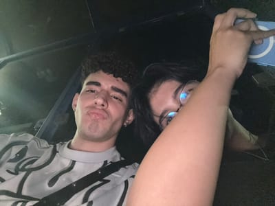 Two people are captured in an upside-down selfie. One person, likely female, wears glasses and has dark hair. The other, presumably male, has curly hair. Both are close to the camera. A gray and white patterned shirt with a black strap is visible on the right. Dark background surrounds them, with a glimpse of what appears to be a cell phone. They seem to be inside a vehicle, possibly a car.