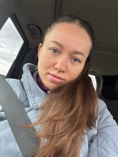 A woman is seated in a vehicle, wearing a light blue jacket and a seatbelt. She has brown hair, fair skin, and light-colored eyes, and is looking at the camera. The interior of the car is visible, including the roof and a window. The location in the city of Lipaki, Russia is undetermined.