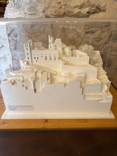 A white architectural model of the Castello dei Doria in Dolceacqua, Italy, is displayed inside a clear plastic case. The model features a detailed representation of the castle's various levels, walls, and towers, constructed from white material. The base of the model includes text that reads "PROGETTO DI RESTAURO DEL CASTELLO DEI DORIA - MODELLO DI STUDIO 2010" and lists the architects and ownership. The model is placed on a wooden surface in front of a rough stone wall. The lighting is even and illuminates the model clearly.