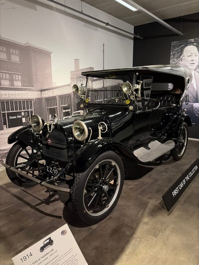 This media file presents an indoor museum display featuring a well-preserved black 1914 Dodge Type 30 Touring Car. The vintage vehicle is polished and equipped with prominent headlights, a front grille, and wooden spoked wheels, with a spare tire mounted on the side. A license plate displaying "ZZ-10" is visible at the front.
The setting includes a dark, mottled floor and walls that transition between dark and light sections, along with overhead metal railings and lighting. In the background, a large black and white mural depicts an industrial building, identified by "DODGE BROTHERS" and "DODGE" signage, likely representing an early factory or dealership. Above this mural, a black and white portrait of a man, possibly Horace Dodge, is displayed.
An overhead black sign highlights the car's significance, stating "FIRST CAR OF THE COLLECTION." A white informational plaque next to the car provides further details, including "1914," "TYPE 30 TOURING CAR," and "FOUR-CYLINDER POWER: 35 HP." The text also mentions "HORACE ELGIN DODGE" and "Dodge gekocht door P.W. Louwman als eerste Louwman Dodge- en Chrysler-importeur en..." which contextually places the exhibit at the Louwman Museum in Wassenaar, Netherlands, celebrating automotive history. The overall scene conveys a sense of historical preservation and appreciation for classic automobiles.