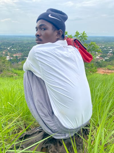 A young Black male wearing a black Nike beanie, a white button-down shirt, and gray sweatpants is crouched on a rock at the top of a hill overlooking a landscape. A bright red purse is slung over his right shoulder. The background shows a sprawling village with red-roofed houses nestled amongst lush green trees and vegetation under a cloudy sky. The foreground consists of tall, vibrant green grass. The location is identified as Ufu Esioni, Nigeria.