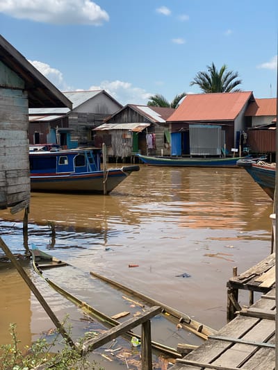This image depicts a view of the village of Berangas Timur, Indonesia, centered on a murky brown waterway. Wooden stilted houses with corrugated metal and wooden siding line the water's edge, with some showing signs of disrepair. Several boats are visible in the water. In the foreground, a partially submerged wooden boat rests at an angle, filled with water and debris. A larger blue and orange boat is moored in the mid-ground. The sky is blue with scattered white clouds, and a palm tree is visible behind the houses. The overall scene conveys a sense of a rural, water-based community.