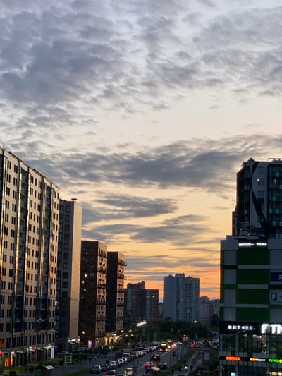 This image depicts a city street scene in Staraya, Russia, during dusk. The sky is filled with a gradient of orange and grey clouds, indicating the setting sun. Several tall residential buildings are visible, with the building on the left being significantly taller and wider than the others. This building features a facade of numerous windows and balconies, reflecting the ambient light. To the right, another modern building displays green and white checkered panels and illuminated signage. One sign, written in Cyrillic script, reads "ВКУСНО - ТОЧКА" (Vkusno - Tochka), which translates to "Tasty - Point" and is a fast-food restaurant chain. Another prominent sign below reads "ФИТНЕС" (Fitness).

The street below is lined with cars, suggesting moderate traffic. Pedestrians are also present on the sidewalks, some of whom are walking towards the buildings. Streetlights are illuminated, casting a warm glow on the street and the lower levels of the buildings. The scene appears to be a busy urban area with a mix of residential and commercial structures.