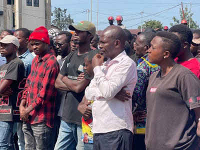 A group of people, primarily men, stand in a line on a street in the city of Ville, Democratic Republic of Congo. The people are wearing a variety of clothing, including shirts, pants, hats, and a few are wearing traditional African attire. The people are all looking in the same direction, but there is no clear focus. The background is composed of a variety of buildings, a cloudy sky, and electric wires. The environment appears to be urban and busy. The image was taken during the day and the lighting is bright. One individual is wearing a red and black plaid shirt. Another person is wearing a black shirt with the word "MARQUE" on it.