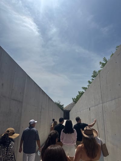A group of approximately ten individuals is depicted walking away from the viewer along a narrow outdoor concrete corridor under a bright, partly cloudy sky. The corridor features tall, unadorned light-grey concrete walls on both the left and right sides, exhibiting visible formwork marks and horizontal seams. Green vegetation is visible over the top edges of both walls. At the far end of the corridor, a structure with a thatched roof and a dark entrance opening is visible. The scene is located in San Isidro Llano Grande, Mexico. Subjects include a person in the foreground left wearing a patterned top and a straw hat; a white male in a white baseball cap, striped polo shirt, and white pants, holding a clear water bottle; a female in a light-colored top and white shorts; and a female in the foreground right wearing a cowboy-style hat and carrying a tote bag. One male subject in a dark shirt, positioned on the right side of the group, has his right arm slightly extended, appearing to touch or gesture towards the right concrete wall.