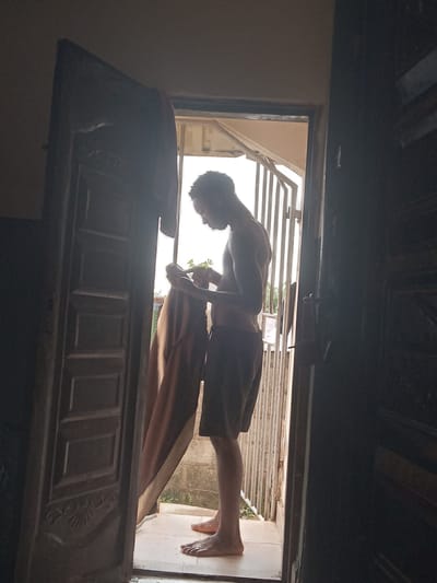 A shirtless person in shorts is standing in the doorway of a house in Achara Ndiagu Omegio, Nigeria. The person is bent over, possibly writing or drawing on something. The door is partially open. There is a gate visible outside the door. The location appears to be residential.