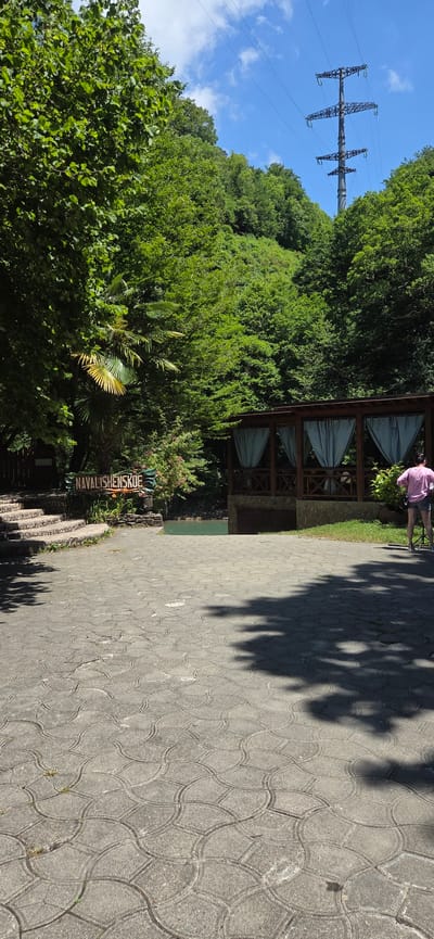 The image depicts a paved outdoor area with a stone walkway leading towards a body of water. To the left, a set of stone stairs ascends, partially shaded by lush green trees. A wooden sign reading "NAVALISHENSKOE" is positioned near the stairs. In the background, a wooden gazebo with blue curtains stands beside the tranquil water. A lone figure wearing a pink shirt and shorts stands facing away from the camera, looking towards the gazebo. The environment is characterized by abundant greenery, suggesting a natural, possibly park-like setting. The sky is bright blue with scattered clouds, and a large electricity pylon is visible in the upper right quadrant of the image, indicating proximity to infrastructure. The paved ground features a repeating interlocking pattern of grey concrete tiles, with shadows of trees cast upon it. The location is identified as Khleborob, Russia.
