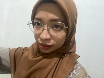 The media file is a close-up, vertically oriented selfie of a young woman wearing a brown hijab and glasses. Her eyes are dark and she is wearing red lipstick. The background is a plain, textured white wall. The lighting appears to be indoor. There is a hint of a garment with embroidery visible on the upper left portion of the image. The overall mood is casual and personal, typical of a selfie. The location context provided is Sekar Mawar, Indonesia.