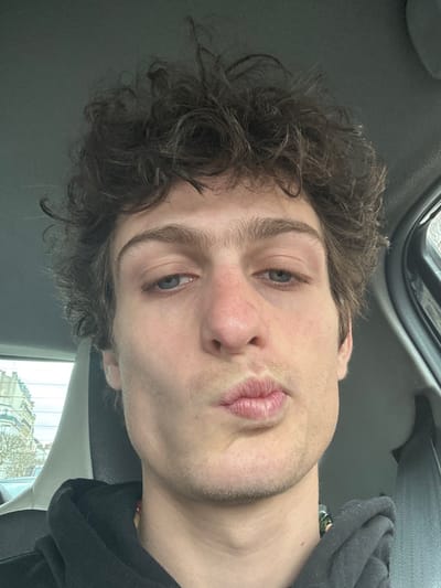 The image captures a close-up portrait of a young adult male inside a vehicle. The subject has short, curly, dark brown hair and blue eyes, which are directed slightly downward and to the viewer's left. His lips are pursed in a kissing expression, and he displays light stubble on his chin and upper lip. He is wearing a black hooded sweatshirt, with a segment of a multicolored beaded necklace visible at the base of his neck on the right. Behind him, the light grey headrest and a portion of the vehicle's interior are visible. Through the rear window on the left side of the frame, distinct Parisian-style architectural facades of stone buildings with multiple windows are visible, alongside bare tree branches under an overcast sky. This urban setting is consistent with the city of Saint-Mandé, France.