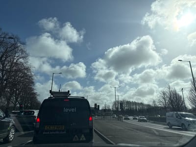 This image depicts a multi-lane arterial road in South Ribble, United Kingdom, under a bright, partly cloudy sky. The scene is captured from inside a vehicle, likely stationary, looking forward.
In the immediate foreground, the rear of a dark grey/black Volkswagen Caddy panel van, registered "CK72 NKD" (UK), is prominently featured. The van displays "level" branding, the website "www.levelltd.com", and two yellow/black triangular hazard symbols (likely indicating hazardous materials). A roof rack on the van carries specialized equipment, including a mast or antenna and a box-like unit, suggesting surveying or telecommunications operations. Its brake lights are illuminated.
To the left of the 'level' van, part of a dark-colored SUV is visible in an adjacent lane. Behind the 'level' van, at least two other vehicles are discernible, also stationary. Traffic lights are visible on both the left and right sides of the road, displaying a red signal for the direction of travel, indicating traffic is stopped.
On the right side of the road, a white panel van is seen traveling in what appears to be an oncoming lane or further down the road. The road surface is asphalt. Tall, dark streetlights line both sides of the roadway.
The environment includes a dense line of bare deciduous trees along the left side of the road, extending into the distance. Additional trees or dense foliage form a treeline in the background on the right. The sky is bright blue, scattered with numerous white cumulus clouds. The sun is visible in the upper right quadrant, casting a strong glare. The overall setting is characteristic of a main thoroughfare or principal road, possibly bordering commercial or light industrial areas within the South Ribble district.