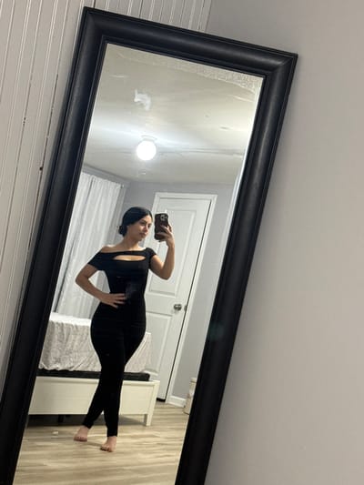 A person stands in front of a large, angled rectangular mirror with a black frame, taking a selfie with a phone. The person is wearing a fitted black outfit with cutouts. Reflected in the mirror is a room with white wainscoting on one wall, a closed white door, and a bed with a patterned comforter. A white bedside table is also visible. A spherical light fixture is reflected on the wall above. The room appears to be brightly lit.