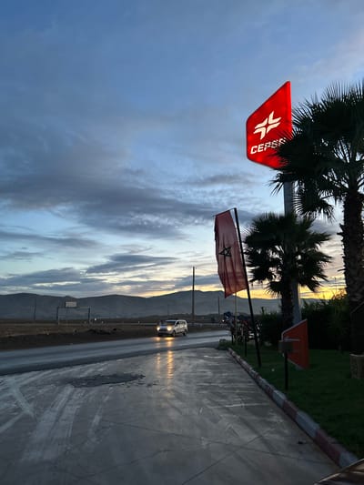 The image displays a serene roadside scene at either dawn or dusk, characterized by a dynamic sky and an illuminated car. The sky dominates the upper portion of the frame, showcasing layers of streaky white and grey clouds that hint at a recent rain shower or atmospheric moisture. A warm, golden light emanating from the horizon suggests the sun is either rising or setting, casting a gentle glow on the distant elements.
In the foreground, a wet, paved road extends towards the right, reflecting the ambient light and the bright headlights of a white or light-colored car driving towards the viewer. A concrete curb with distinctive red and white stripes runs alongside the road. To the left of the road, a red, triangular sign bearing the white "CEPSA" logo is prominently displayed, alongside a Moroccan flag, identifiable by its red field and green pentagram. Several palm trees with dark, feathery fronds stand nearby, their forms silhouetted against the cloudy sky. Further down the road, a small orange directional sign is partially visible.
The mid-ground and background feature a range of mountains, their silhouettes softened by the atmospheric conditions and the distant light. The overall scene is devoid of people, emphasizing the natural landscape and the quiet presence of infrastructure. The visible Moroccan flag and CEPSA sign, combined with the landscape of palm trees and mountains, strongly indicate a location in Morocco, consistent with Ain Kansra.