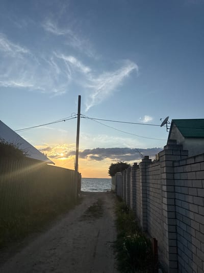 A dirt path leads towards the sea, bordered on the left by a corrugated metal fence and on the right by a brick wall. The sun is low in the sky, casting a warm glow and creating visible light rays. The sky is blue with scattered white clouds. A utility pole with wires stands in the middle of the path. The sea is visible at the end of the path. The overall scene suggests a coastal area in Zatoka, Ukraine.