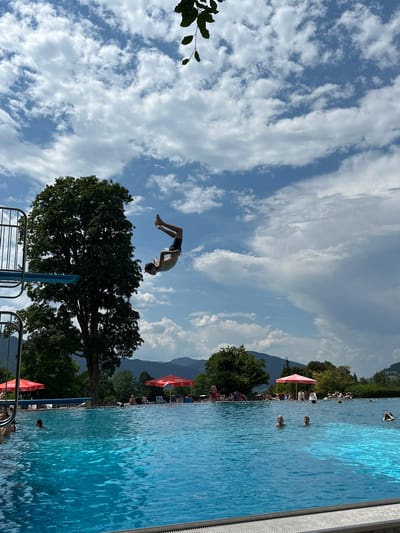 A young male is captured mid-air performing a flip off a diving board at an outdoor swimming pool in Oberammergau, Germany. The diving board is on the left of the frame, with a tall, lush green tree behind it. The pool itself is a vibrant blue, with several people swimming and relaxing in and around it. Red umbrellas provide shade on the deck. In the background, rolling green hills and mountains are visible under a partly cloudy blue sky. The scene conveys a sense of summer recreation and activity.