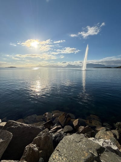 A large water fountain is erupting in the distance, shooting a tall plume of water into the sky. The fountain is located in the sea, with landmasses and mountains visible on the horizon. The sun is shining brightly, casting a glare on the water and creating reflections. The foreground of the image is lined with large rocks, forming a natural barrier along the water's edge. The sky is blue with scattered clouds. The scene appears to be set in Harstad, Norway, a coastal city known for its natural beauty.
