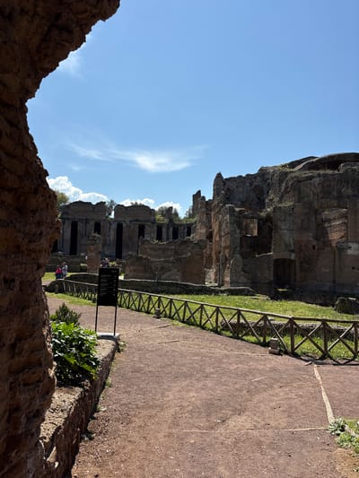 The image depicts a scene at Hadrian's Villa, located in Tivoli, Italy. The ruins of an ancient structure stand prominently in the background, with several visible arched openings and varying levels of decay. A wooden fence runs alongside a dirt path, and a signpost with directional information is positioned near the path. Two individuals, possibly tourists, are situated in the middle ground. In the foreground, a section of a stone wall frames the lower part of the view, adding depth and perspective to the scene. The sky above is clear with scattered clouds.