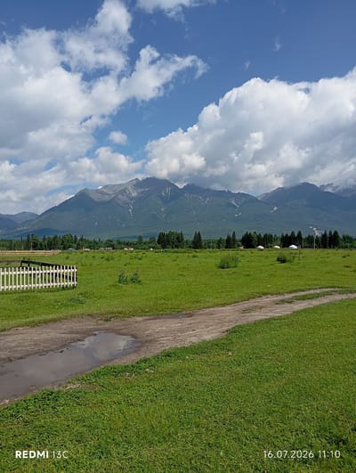 A wide-angle landscape photograph depicts a rural scene with a dirt path leading through a green grassy field. In the foreground, a puddle reflects the sky. A white picket fence is visible on the left side of the frame. In the middle ground, several white yurts are scattered across the field, and a line of trees forms a dark green border. In the background, a majestic mountain range rises, its peaks partially obscured by clouds. The sky is a vibrant blue, dotted with fluffy white clouds. The bottom of the image displays text indicating "REDMI 13C" and the date "16.07.2026 11:10". The location is identified as Khurai-Khobok, Russia.