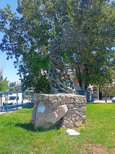 A metallic sculpture of a stylized figure with outstretched wings stands on a stone pedestal in a grassy area in Chalkida, Greece. The sculpture appears to be made of twisted metal or wire, giving it a skeletal and ethereal quality. Behind the sculpture, a large, mature tree with dense green foliage dominates the background, with its branches reaching towards a clear blue sky. To the left of the sculpture, a white van is parked under a blue awning structure, suggesting a public area such as a park or promenade. In the distance, buildings and more trees are visible, indicating an urban or semi-urban setting. The lighting suggests a bright, sunny day. A small white plaque is attached to the stone base of the sculpture.