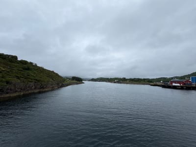 The image shows a wide, calm body of water, likely a fjord or bay, under an overcast sky. On the left, a steep, green, and rocky hillside slopes down towards the water. The right side of the image features a small settlement with several red and white buildings, some appearing to be houses and others possibly industrial or commercial structures, situated along a dock. A few boats are visible tied up at the dock. The water is dark and slightly rippled. The overall impression is of a quiet, possibly remote coastal area.