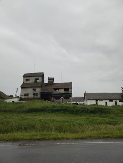 The image depicts a rural scene in Lakly, Russia, under an overcast sky. The primary subjects are a cluster of industrial or agricultural buildings situated on a gentle slope. The most prominent structure is a multi-story building with a corrugated metal exterior, appearing weathered and aged. It has several windows, some of which are broken. Adjacent to this main building are several smaller, white-painted structures with corrugated roofs, resembling farm outbuildings or barns. The foreground features a lush, green grassy field, and the very bottom of the image shows a portion of a wet asphalt road, suggesting recent rain. The overall atmosphere is one of a rural, possibly disused, industrial or agricultural site. The time of day appears to be daytime, given the ambient light, but the cloudy weather creates a muted, somber mood. There is no discernible text in the image. The scene suggests a quiet, potentially isolated location in the Russian countryside.