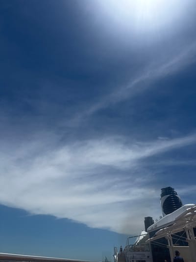 A bright sun illuminates a deep blue sky dotted with cirrus clouds stretching across the frame. In the upper right corner, a partial view of a cruise ship reveals its upper decks and some passengers. A metal railing runs along the bottom edge of the picture, providing a sense of perspective. The sky dominates the scene, with the ship appearing as a minor element against the vast expanse above.