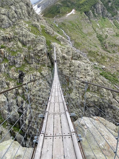 This photograph captures a first-person perspective from the beginning of a narrow, wooden suspension bridge spanning a deep, rocky gorge in Innertkirchen, Switzerland. The bridge, constructed of light-colored wooden planks secured by dark circular bolts, extends diagonally from the lower left to the center, leading into the distance. Heavy-gauge braided metal cables form the primary suspension lines, running along both sides of the bridge, supported by a network of thinner vertical and diagonal metal wires that serve as railings and safety barriers. These wires are affixed to the main cables and bridge structure with metal clamps and rings.

One human figure, appearing as a small dark silhouette, is visible on the bridge deck in the upper middle distance. The surrounding environment consists of steep, rugged mountainsides composed of gray, weathered rock formations with extensive fracturing. Patches of green vegetation, including moss, grass, and shrubs, adhere to the rocky surfaces. In the upper right quadrant, a thin stream of water cascades down the mountainside, disappearing into the lower terrain. Further up the distant mountains, patches of white snow are visible, indicating higher elevation. The sky above is overcast, presenting a diffused light. The immediate foreground on the right shows the edge of a large, light-colored rock outcrop partially covered with lichen, adjacent to the bridge's structural elements.