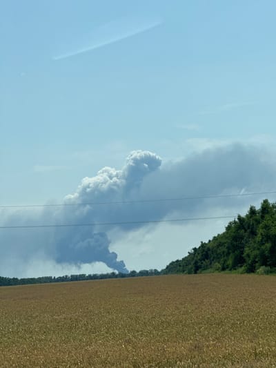 The image depicts a rural landscape with a field of wheat in the foreground under a clear blue sky. In the middle ground, a dense line of green trees runs along a gentle slope. In the background, a large plume of dark smoke rises into the sky, suggesting a fire or industrial activity. Two power lines are visible in the middle distance, stretching horizontally across the frame. The scene is observed from a distance, with no people or specific structures immediately identifiable. The overall impression is one of a tranquil natural setting disrupted by an external event indicated by the smoke plume. There is no specific indication of the location being within the city of Рождественская, Russia; it appears to be a rural area.
