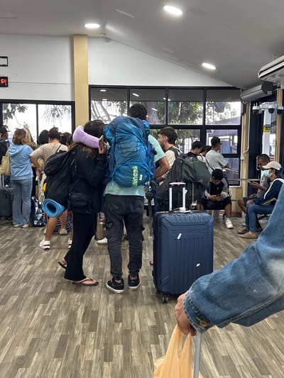 The image depicts a busy indoor waiting area, likely a ferry terminal or bus station, in Tambon Don Sak, Thailand. A diverse group of people, predominantly tourists, are gathered, many with backpacks and luggage. The atmosphere is one of transit and anticipation.
The environment features a light-colored tiled floor with a wood-grain pattern. Large windows with dark blinds line the back wall, revealing glimpses of outdoor greenery and trees. Fluorescent lighting fixtures are mounted on the angled ceiling. A digital clock displaying "21:54" is visible on the upper left wall. Several CCTV cameras are mounted on the ceiling and walls, suggesting a security-conscious environment.
Key subjects include:
* **People:** A variety of individuals are standing, waiting, and moving through the space. Many are dressed casually in t-shirts, shorts, and jeans. Several individuals are wearing backpacks, indicating travel. Some are looking at their phones, while others are engaged in conversation.
* **Objects:**
* **Luggage:** Several large suitcases, including a prominent navy blue "TROOPER" suitcase in the foreground, are present. Backpacks of various sizes and colors are worn by many individuals.
* **Bags:** Numerous travel bags, tote bags, and plastic bags are visible. One person in the foreground is holding a light orange plastic bag.
* **Other:** A blue yoga mat is strapped to a backpack. A purple travel pillow is being held by a woman.
Actions and interactions: People are primarily standing and waiting in queues or scattered groups. Some appear to be checking schedules or communicating with travel companions. The overall impression is one of organized chaos associated with travel hubs. The presence of luggage and the crowded nature of the scene suggest that a ferry or bus departure is imminent.