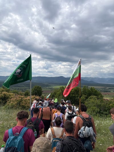 A high-angle view captures a large group of people seated on a hillside in Mramoren, Bulgaria, facing away from the camera. Visible are the backs of their heads and various backpacks. A green flag with white lettering in Bulgarian and a white edelweiss logo stands on the left, while a red, white, and green horizontal-striped flag with Bulgarian text and a similar logo stands further away on the right. Beyond the group and flags, a landscape featuring trees, mountains, and a cloudy sky stretches into the distance. The hillside appears to be covered with grass and small white flowers.