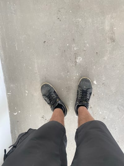 The image shows a pair of black, worn sneakers and black shorts on a concrete surface. The sneakers have black laces and yellow accents. The concrete appears to be outdoors and is speckled with small white and dark marks, suggesting it may be under construction or in an industrial area. The lighting indicates it is daytime. The perspective is from above, looking down at the person's legs and feet. The scene does not provide any specific indicators that would allow for a precise location in the city of Breda, Netherlands.
