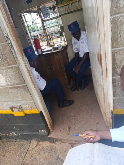 The image depicts a scene inside and immediately outside a small security booth in Kaplelwo, Kenya. Two security guards are seated within the cramped booth, viewed through its open doorway. Both guards are uniformly dressed in blue caps displaying a "PRECISION SECURITY" logo, white short-sleeved shirts with a similar company emblem, dark blue trousers, and black shoes. The guard on the right looks towards the camera, while the one on the left looks downwards.
Inside the booth, a simple wooden desk or cabinet holds a red thermos bottle and two white mugs. Behind the guards, a barred window allows natural light to enter and offers a glimpse of the outdoor environment, including greenery and distant structures. A calendar, showing "APRIL", "MAY", and "JUNE" along with numerical dates, is affixed to the wall above the desk.
In the foreground, partially visible, a third individual stands outside the booth. This person is holding a blue and yellow pen over an open notebook or ledger filled with handwritten text, suggesting they are taking notes, conducting a check, or interacting with the guards for a report.
The ground outside the booth is concrete, and the lower exterior walls of the booth are painted dark with a yellow stripe. The scene is illuminated by natural daylight, indicating it is daytime. The overall impression is of a routine administrative or reporting activity at a security post.
