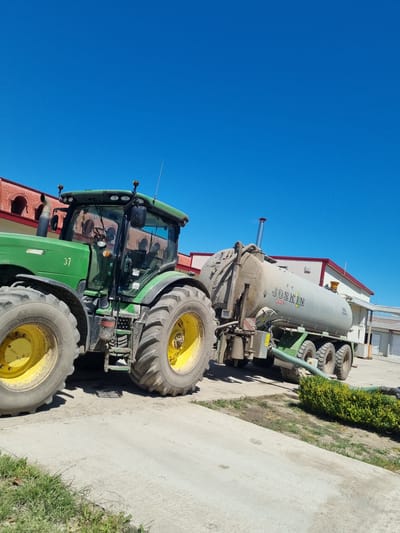 The image shows a green John Deere tractor coupled to a Joskin manure spreader, both parked on a concrete surface in an outdoor setting. The sky is clear and blue. In the background are buildings with red-trimmed roofs and white walls, and a hedge is visible along the side of the concrete surface. Based on the prompt, the location is Ovcharovo, Bulgaria.