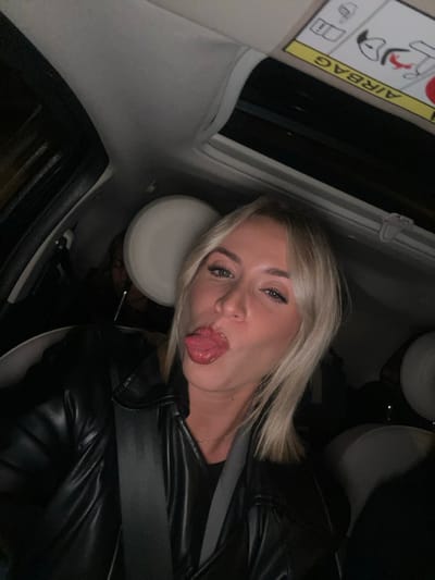 A blonde woman is in a car, sticking her tongue out at the camera. She is wearing a black leather jacket and seatbelt. The car interior is visible, including the airbag label and headrest. The image is taken from an unusual angle, suggesting the camera is positioned overhead.