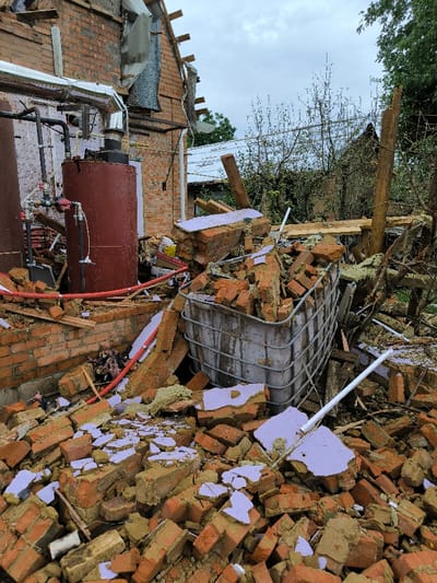 The image displays a scene of significant structural damage in Pysarivka, Ukraine. The foreground is covered with an extensive accumulation of broken reddish-brown bricks and fragments of light purple insulation material. A large, light-colored plastic intermediate bulk container (IBC) enclosed within a metal cage is positioned towards the center-right, partially collapsed and filled with bricks and debris. To the left, a brick building exhibits substantial damage to its upper section, with exposed wooden rafters and fragmented roofing elements visible. Affixed to the exterior brick wall of this building are two large cylindrical tanks, one dark red and one dark brown, connected by a network of silver-colored insulated pipes, red flexible tubing, and valves. Additional scattered debris, including sections of red pipe, lies on the ground around the tanks. The background reveals sparse trees and vegetation beneath an overcast sky. No individuals are present within the depicted scene.