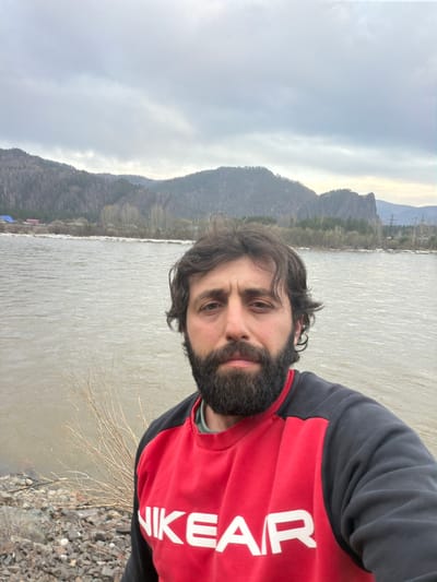 A self-portrait captures a man outdoors, positioned on the right side of the frame. He has dark, wavy hair and a full beard, looking directly at the camera with a neutral expression. He is wearing a two-tone sweatshirt, primarily red on the chest and black on the sleeves and shoulders. White text spelling "NIKE AIR" is clearly visible across the red section of his sweatshirt.
The background features a broad body of water, likely a river, reflecting the overcast sky with a light, rippled surface. Beyond the water, a line of rugged mountains stretches across the horizon, with forested slopes visible in parts. The sky is predominantly grey and cloudy, with patches of brighter light visible near the mountain tops, indicating an overcast day or possibly diffused sunlight during sunrise or sunset. The immediate foreground on the right, beside the man, consists of a rocky shore and some dry, thin branches. The scene conveys a cool, natural environment.