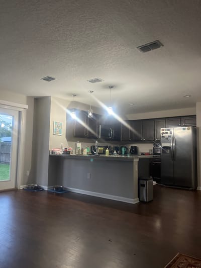 This is a wide-angle, eye-level interior shot of a modern kitchen and dining area. The flooring is dark hardwood. The left side of the image shows a glass door leading to an outdoor area with a grassy lawn and a wooden fence. Inside, near the door, are two pet food bowls on blue mats. The kitchen features dark stained cabinetry, granite countertops, and stainless steel appliances, including a refrigerator, microwave, and dishwasher. A gray island with a granite countertop separates the kitchen from the dining area. Pendant lights hang over the island. The ceiling has textured finish and includes two air vents. A silver trash can is located near the island. A framed picture hangs on the wall near the kitchen. The lighting suggests it is daytime or evening with artificial light. There are no people or animals visible in the image. The location is a residential dwelling in Jacksonville, United States of America.