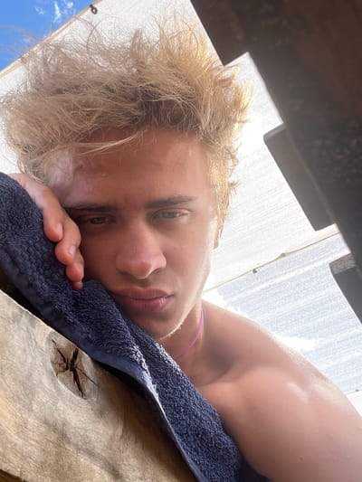 A close-up, low-angle, outdoor selfie captures a young Caucasian male with tousled blond hair leaning his head on a dark blue towel. The man's left arm is visible, resting on a weathered wooden surface with a knot. He is shirtless, wearing a thin purple necklace. His gaze is directed slightly downward and to the left, with a neutral expression. The background consists of a bright, white, fabric-like structure partially obscured by a dark, angular object on the right. Above the structure, a clear blue sky with a few wispy clouds is visible. The lighting suggests a sunny day, with the sun directly behind the man, creating a slight backlight effect and highlighting his skin with a dewy sheen. The location is described as the city of Gres-2, Kazakhstan.
